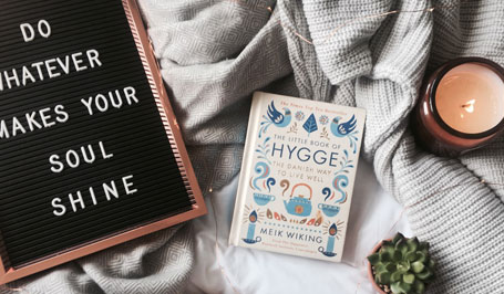 Bett auf dem eine graue Kuscheldecke, ein Schild mit Message, ein Buch zum Thema Hygge und ein Teelicht liegen