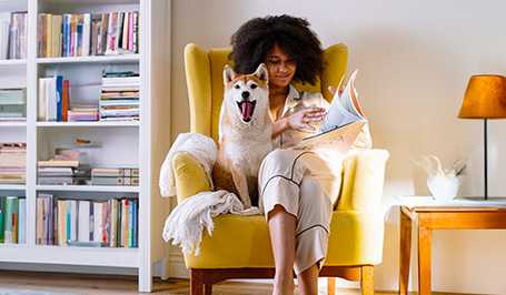 Junge Frau sitzt mit ihrem Hund in einem gelben Ohrensessel und blättert in einem Magazin