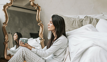Junge Frau sitzt im Pyjama neben ihrem Bett und genießt ihren Kaffee. Ihr Gesicht spiegelt sich in einem Standspiegel