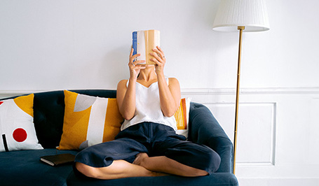 Eine Frau sitzt auf einer dunkelblauen Couch und verdeckt ihr Gesicht mit einem Buch. Neben der Couch steht eine goldene Stehlampe mit weißem Lampenschirm