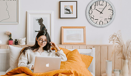 Junge Frau mit Brille sitzt mit einem MacBook im Bett. Hinter ihr hängen an der Wand ein paar Bilder und eine XXL-Wanduhr